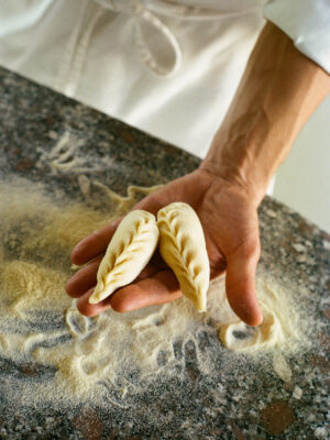 A person holds two neatly braided, uncooked dumplings in their hand over a floured countertop, with flour scattered around and another dumpling resting nearby.