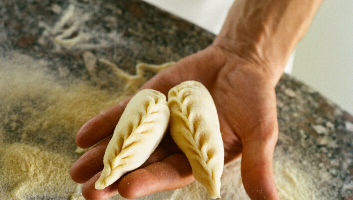 Una persona tiene in mano due paste intricate e intrecciate su un piano di lavoro infarinato. I pasticcini hanno bordi decorativi e la farina è sparsa sulla superficie.