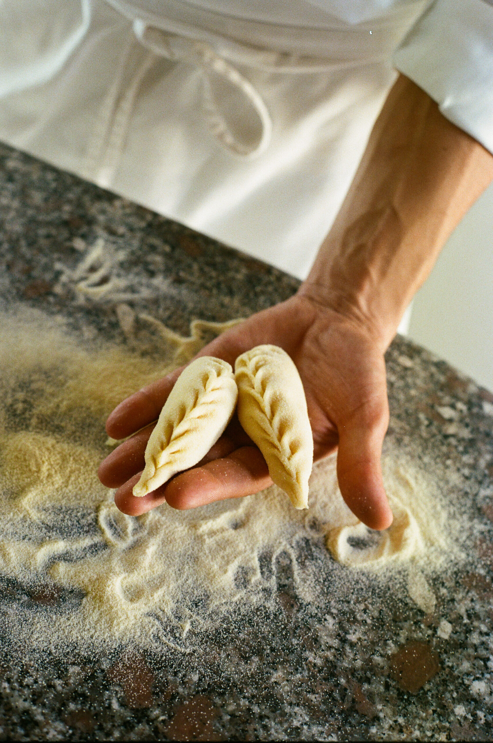 A person holds two neatly braided, uncooked dumplings in their hand over a floured countertop, with flour scattered around and another dumpling resting nearby.