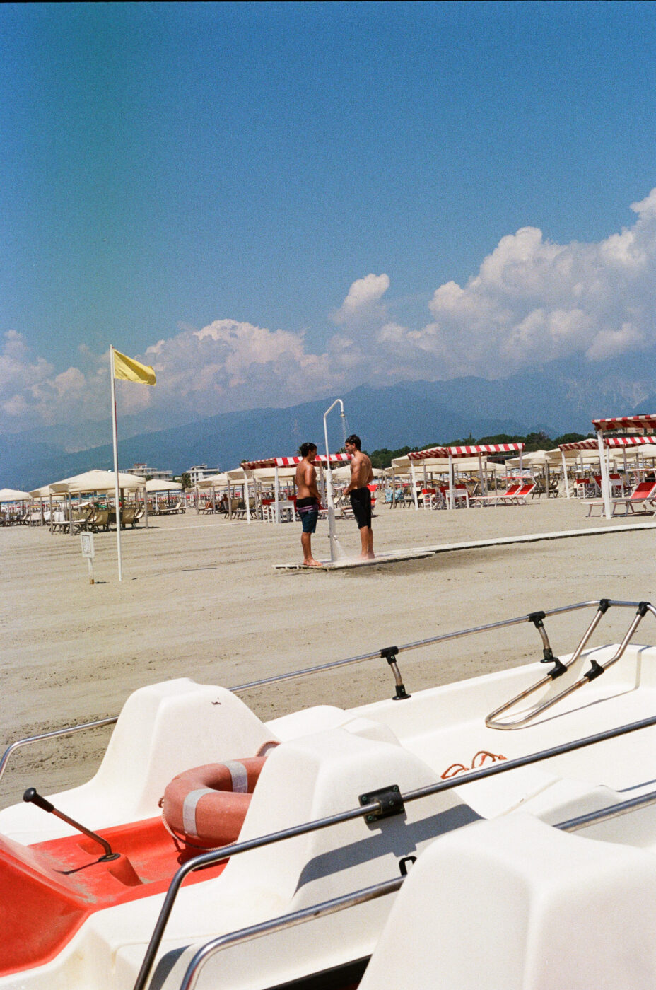 [Due persone si sciacquano in una doccia all'aperto vicino a ombrelloni a strisce bianche e rosse su una spiaggia sabbiosa. In primo piano, è visibile un pedalò bianco e rosso con un anello di salvataggio. Il cielo è parzialmente nuvoloso.