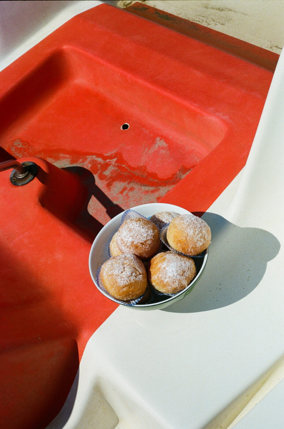 Il titolo dell'immagine non è stato fornito, quindi il testo alt dovrebbe essere: Una ciotola di pasticcini rotondi spolverati di zucchero a velo si trova su una superficie rossa e bianca, forse parte di una barca o di un mobile da esterno, in piena luce solare.