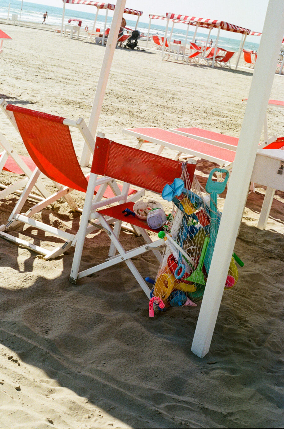 Sedie e ombrelloni rossi - Sedie e ombrelloni rossi su una spiaggia sabbiosa, con una borsa di giocattoli da spiaggia colorati appesi a un palo bianco. L'oceano è visibile sullo sfondo sotto un cielo limpido.