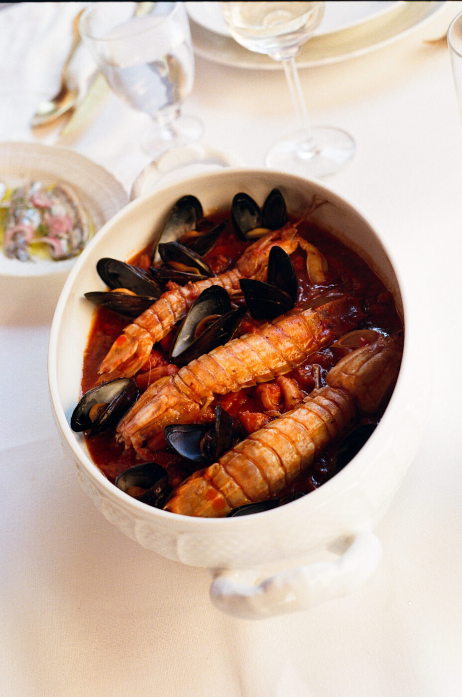 Stufato di frutti di mare - Un piatto da portata bianco riempito di stufato di frutti di mare, con grandi scampi e cozze nere in una ricca salsa di pomodoro rosso, posto su una tovaglia bianca con bicchieri e piatti sullo sfondo.