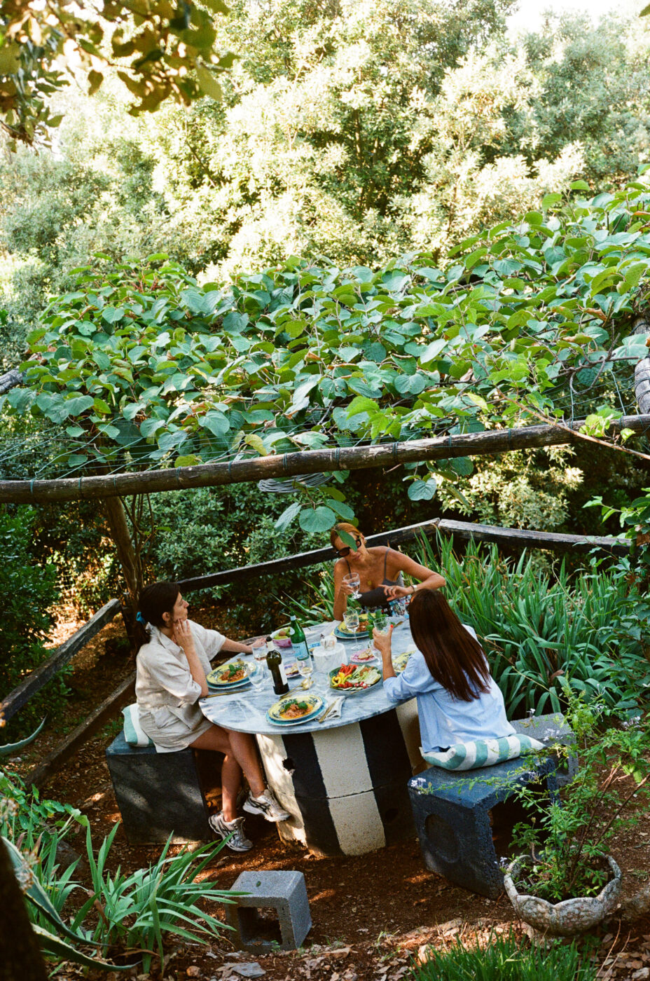 Tre persone a cena all'aperto - Tre persone siedono intorno a un tavolo rotondo di pietra all'aperto, condividendo un pasto tra la vegetazione lussureggiante e una tettoia di foglie in alto. I piatti di cibo e le bevande sono sul tavolo, creando una scena rilassata e naturale.