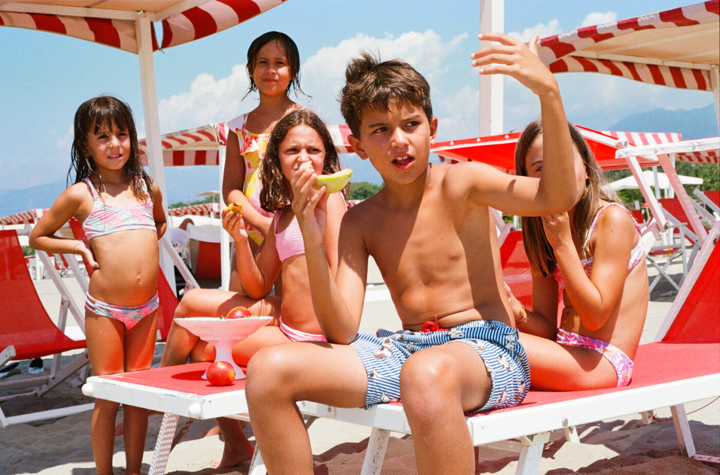 Non è presente il titolo dell'immagine - Cinque bambini in costume da bagno siedono e stanno in piedi accanto a sedie a sdraio e ombrelloni bianchi e rossi su una spiaggia assolata. Alcuni mangiano anguria e un ragazzo in pantaloncini a righe fa dei gesti con il braccio mentre parla al gruppo.