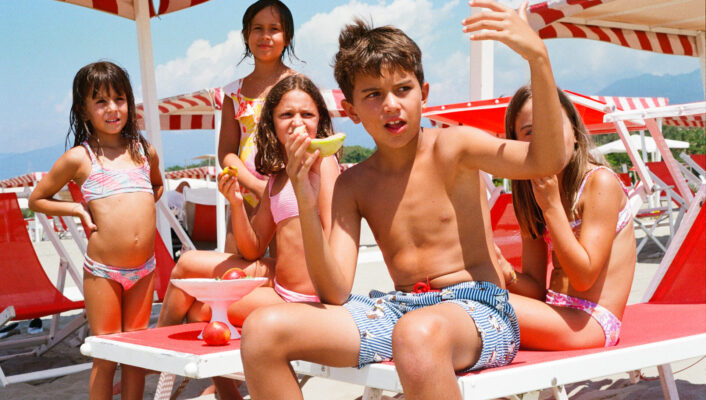 Non è presente il titolo dell'immagine - Cinque bambini in costume da bagno siedono e stanno in piedi accanto a sedie a sdraio e ombrelloni bianchi e rossi su una spiaggia assolata. Alcuni mangiano anguria e un ragazzo in pantaloncini a righe fa dei gesti con il braccio mentre parla al gruppo.