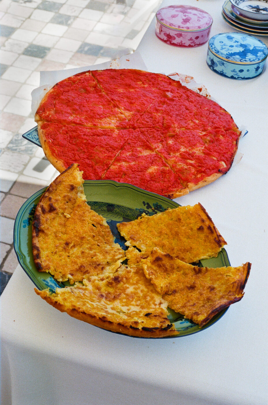 Pizza al pomodoro e focaccia - Su un tavolo sono esposti due piatti: una pizza al pomodoro rotonda e sottile tagliata a fette e una focaccia spezzata e dorata su un piatto verde. Sullo sfondo, lattine decorative e un pavimento a scacchi.