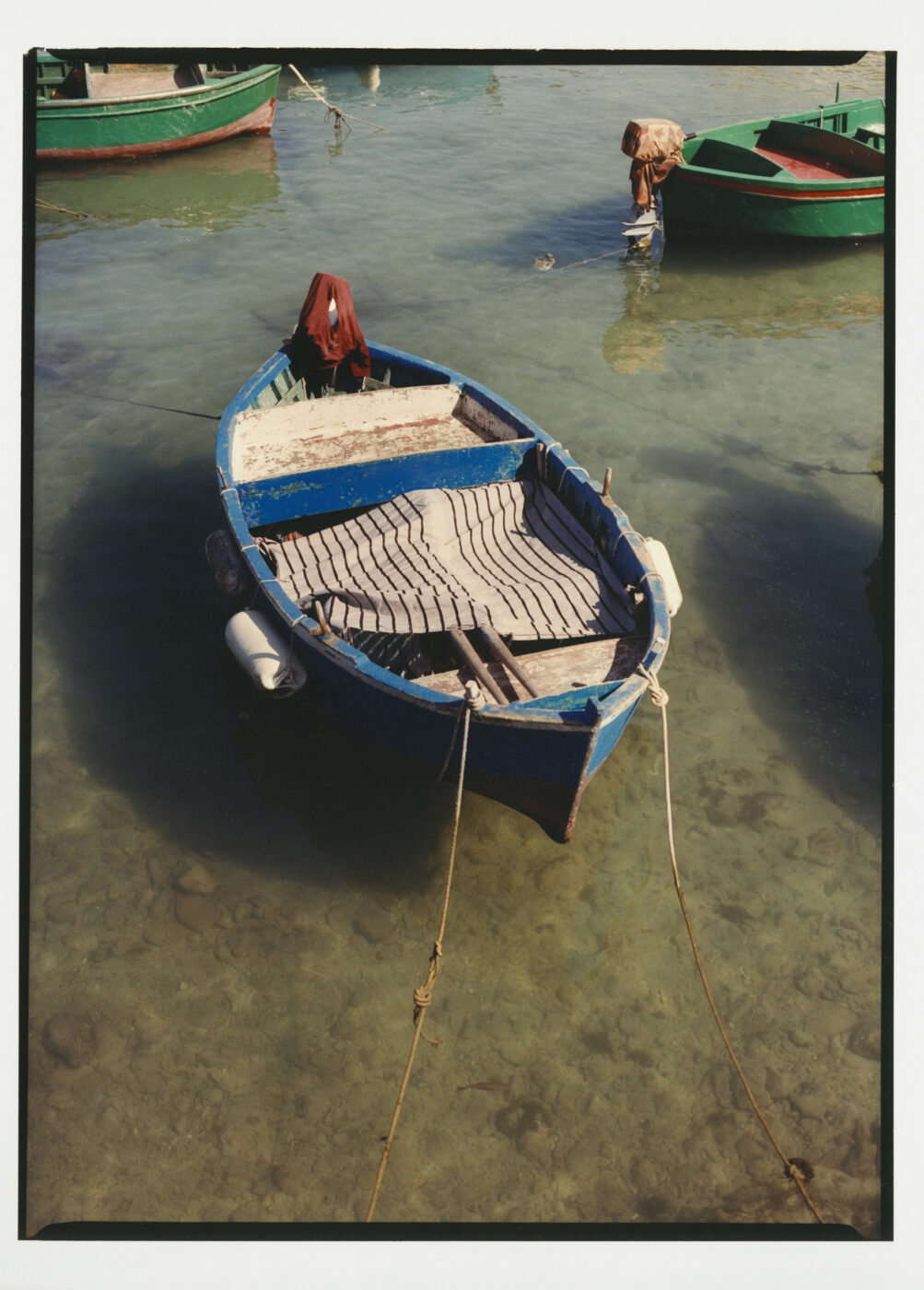 A blue wooden rowboat is anchored in clear, shallow water - A blue wooden rowboat is anchored in clear, shallow water. Two ropes tie the boat to shore, and other green boats are visible nearby. The boats interior appears weathered and empty.