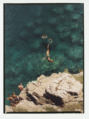 Cliff Diving Adventure - A person dives off a rocky cliff into clear, blue-green water, while three swimmers wait below and two other people stand on the rocks, watching. The scene is bright and summery. If no image title is present, just use: A person dives off a rocky cliff into clear, blue-green water, while three swimmers wait below and two other people stand on the rocks, watching. The scene is bright and summery.