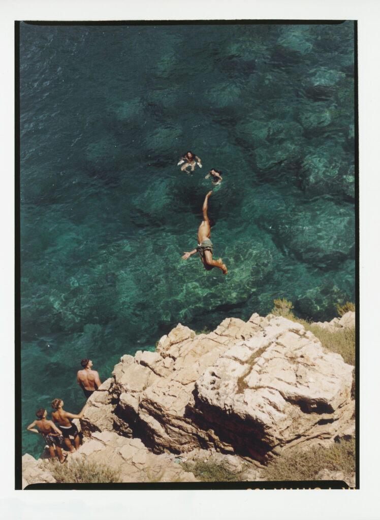 Cliff Diving Adventure - A person dives off a rocky cliff into clear, blue-green water, while three swimmers wait below and two other people stand on the rocks, watching. The scene is bright and summery. If no image title is present, just use: A person dives off a rocky cliff into clear, blue-green water, while three swimmers wait below and two other people stand on the rocks, watching. The scene is bright and summery.