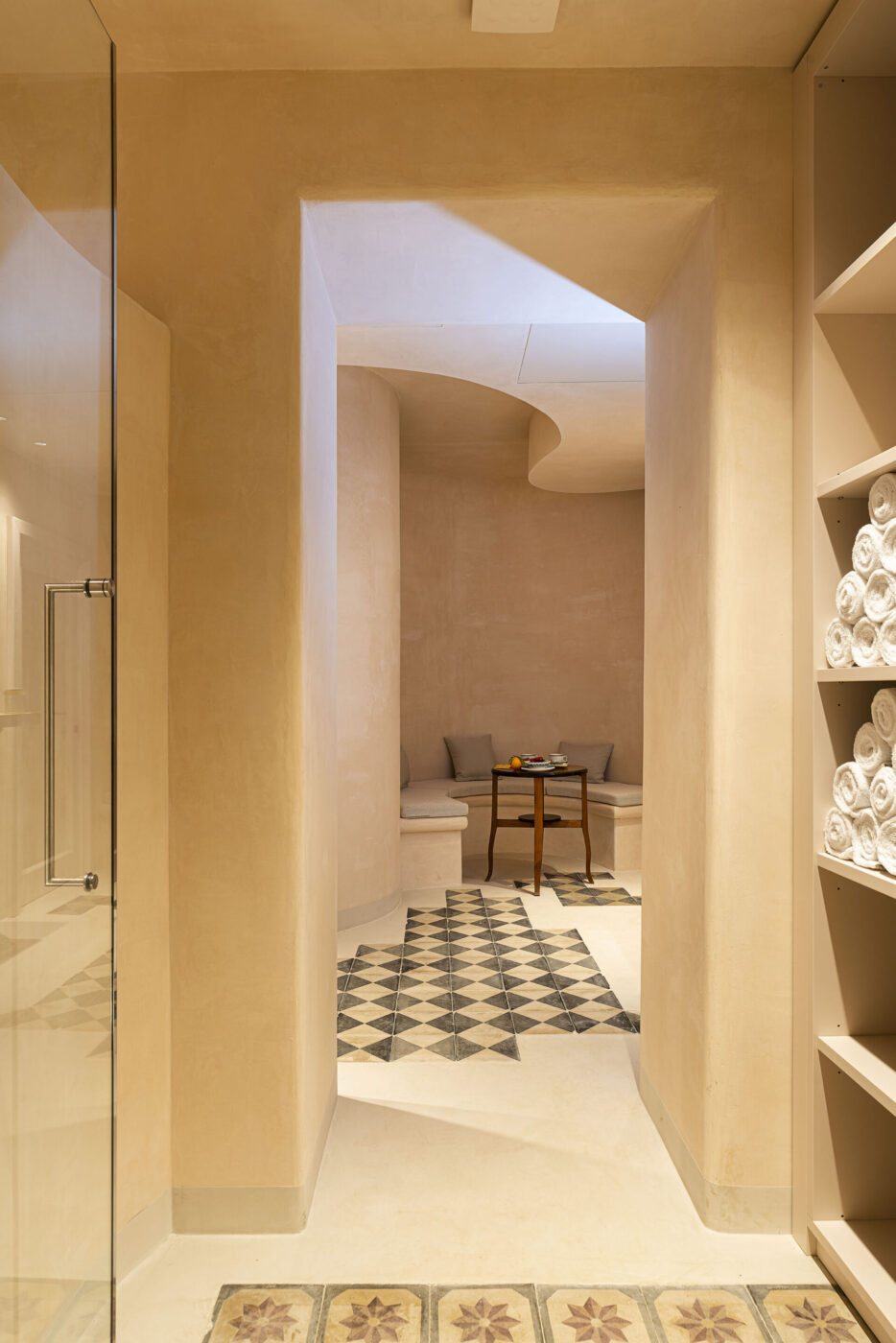 Modern Spa Interior - A modern spa interior with warm beige walls, patterned floor tiles, rolled white towels on shelves, and a cozy seating area with a small wooden table and cushions visible through an open doorway.