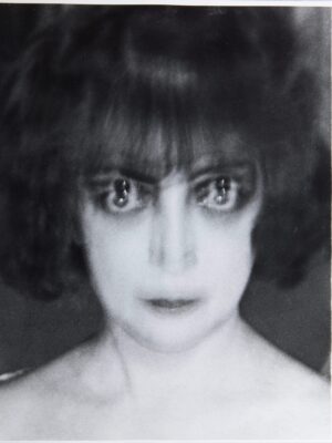 Portrait of: Luisa Casati Stampa A black and white blurry photo of a woman with shoulder-length hair and bangs, appearing to have two sets of eyes due to a double exposure effect. She looks directly at the camera with a blank expression.