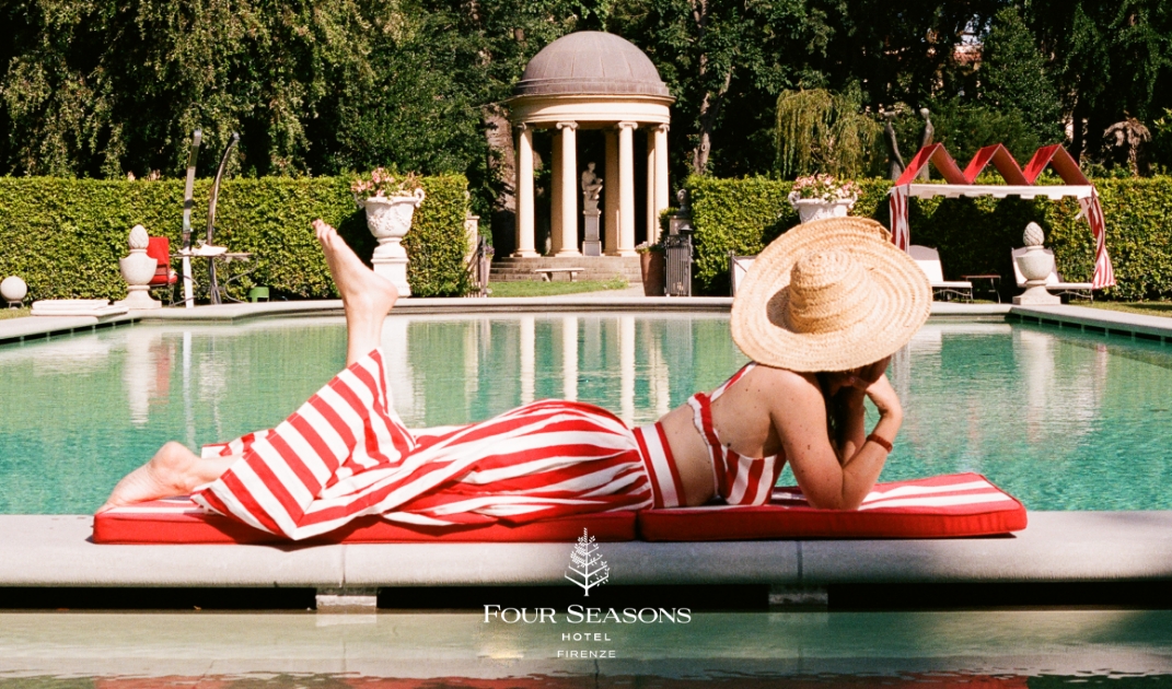 Image Title: Relaxing at Four Seasons Hotel Firenze - A woman in a wide-brimmed hat and red-and-white striped dress lounges on a red mat beside a pool, with gardens and a domed gazebo visible in the background at Four Seasons Hotel Firenze.