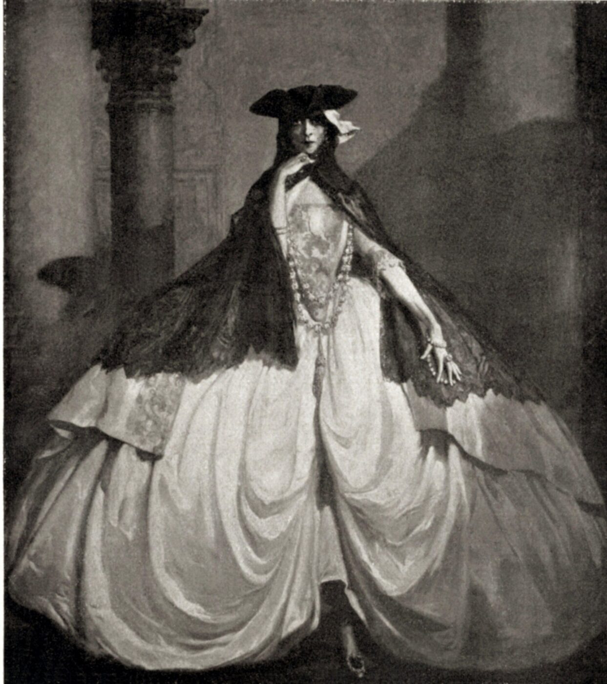 Certamente! Ecco il testo alt, formattato secondo le vostre istruzioni: [Titolo immagine] - Una persona in un elaborato costume storico con un'ampia gonna bianca, dettagli in pizzo, un mantello scuro, un cappello a punta e una maschera si trova in una posa drammatica davanti a un'alta colonna. L'immagine è in bianco e nero.