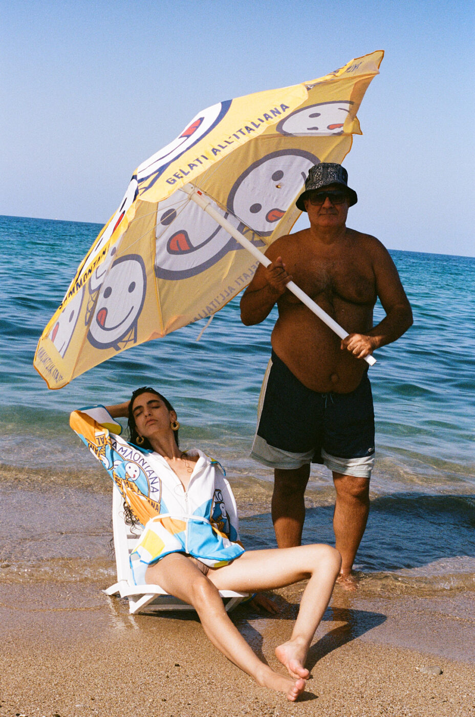 Non è stato fornito il titolo dell'immagine. Se il titolo dell'immagine è, ad esempio, "Beach Relax", il testo alt appropriato sarà: Relax in spiaggia - Una persona si rilassa su una sedia da spiaggia sotto un ombrellone giallo con faccine sorridenti, tenuto da un'altra persona in piedi accanto a lei. Si trovano su una spiaggia sabbiosa con il mare e il cielo blu sullo sfondo. Se avete un titolo dell'immagine da utilizzare, sostituitelo all'inizio secondo il vostro formato!.