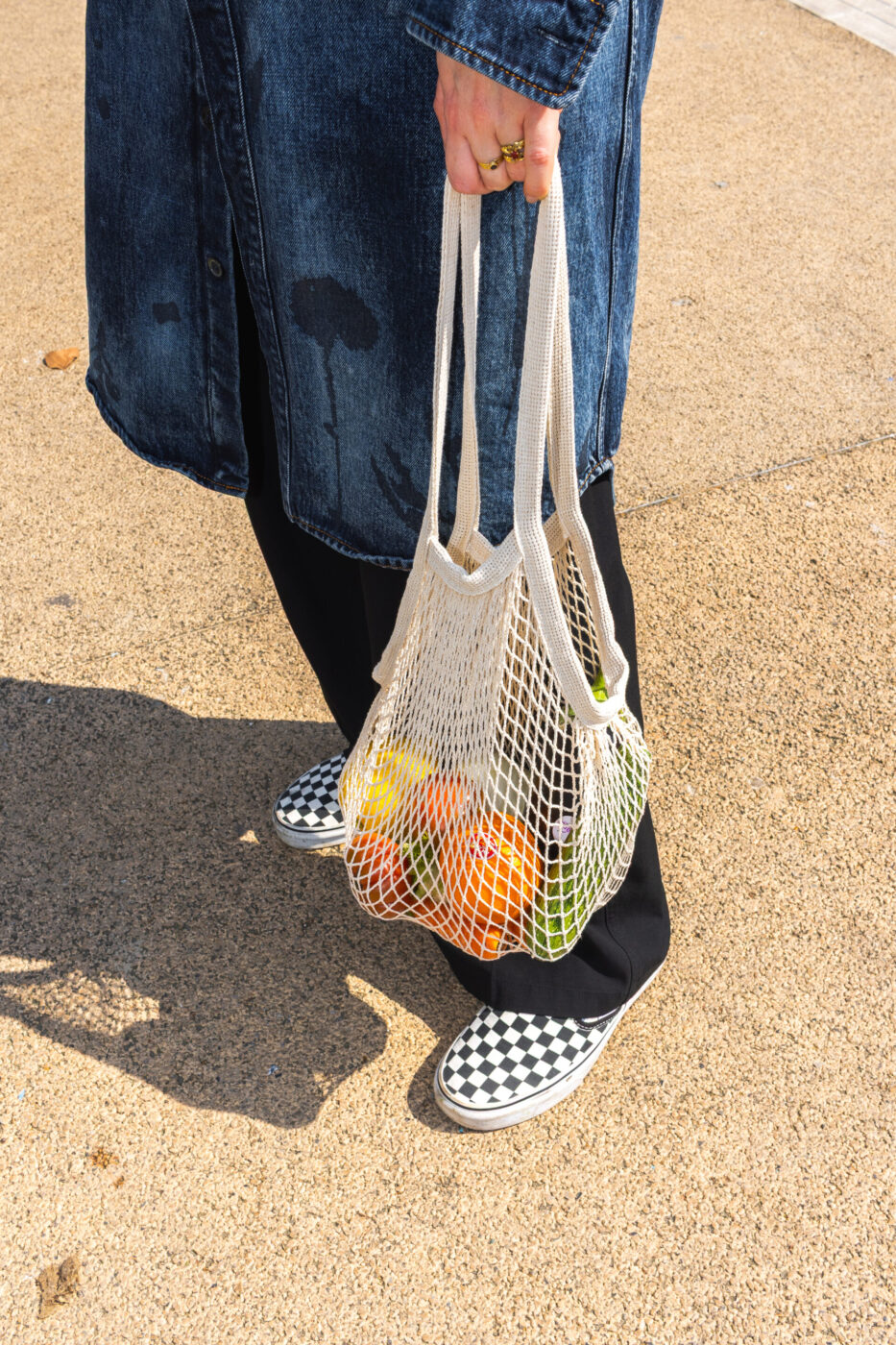 Una persona che indossa un cappotto di jeans blu e pantaloni neri tiene in mano una borsa della spesa in rete con della frutta all'interno. È in piedi su una superficie sabbiosa e indossa scarpe slip-on a scacchi bianchi e neri.