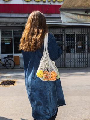 A person with long brown hair, wearing a denim jacket, stands outdoors with their back to the camera, holding a mesh bag containing colorful fruits and vegetables. A storefront and bicycle are visible in the background. (Note: Since you did not provide an image title, the alt text does not include one at the beginning. If you provide an image title, I will prepend it as instructed.).
