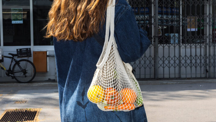 A person with long brown hair, wearing a denim jacket, stands outdoors with their back to the camera, holding a mesh bag containing colorful fruits and vegetables. A storefront and bicycle are visible in the background. (Note: Since you did not provide an image title, the alt text does not include one at the beginning. If you provide an image title, I will prepend it as instructed.).