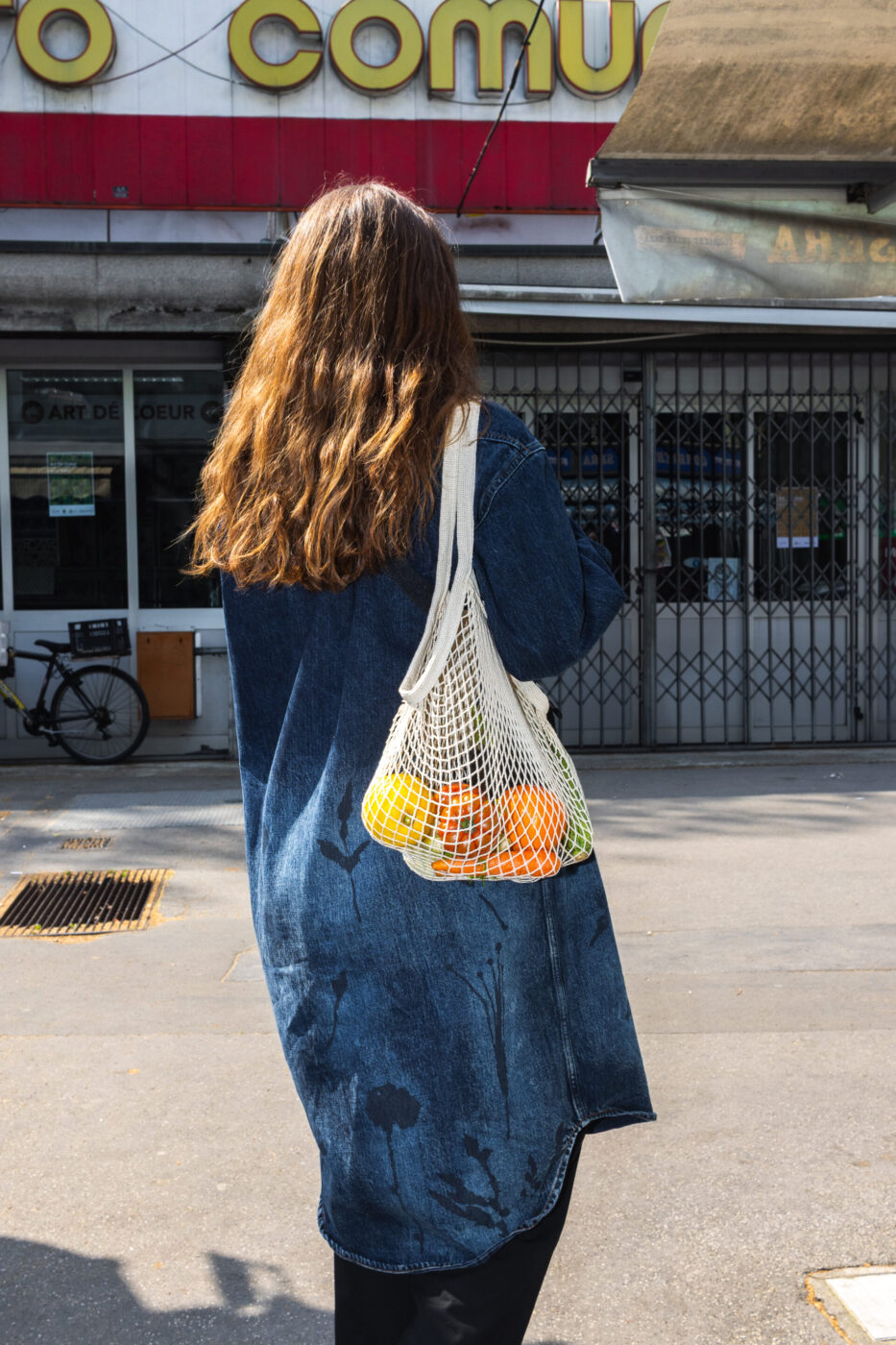 [Una persona con lunghi capelli castani, che indossa una giacca di jeans blu, si trova all'aperto con in mano un sacchetto di rete pieno di arance e limoni. La persona è rivolta verso la telecamera e sullo sfondo è visibile la vetrina di un negozio.