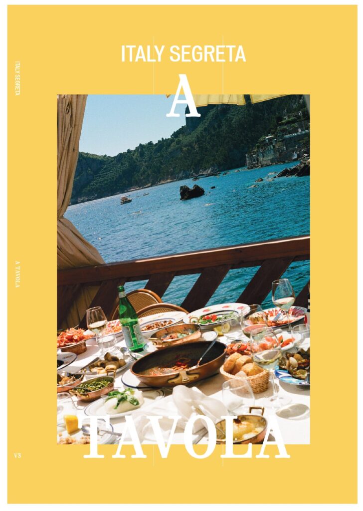 Italy Segreta A Tavola - A sunlit table set with a variety of Italian dishes and drinks overlooks blue water and rocky hills, viewed from a shaded outdoor area. Text reads Italy Segreta A Tavola.