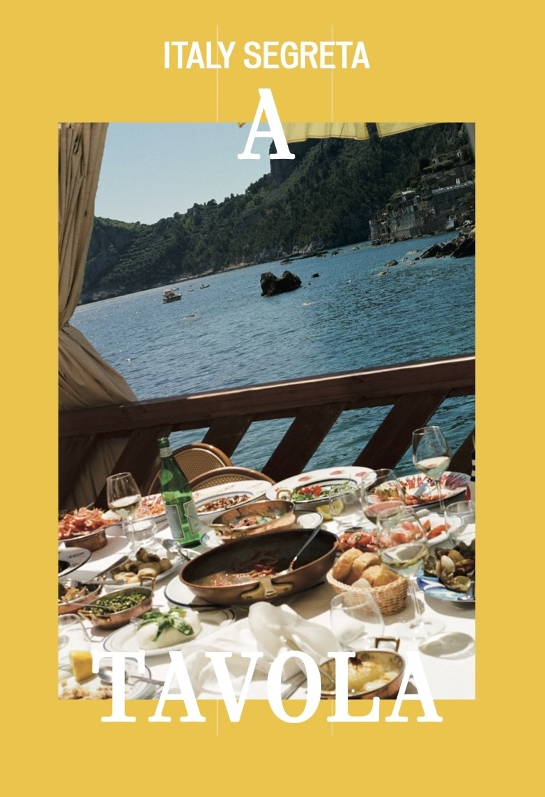 A TAVOLA (2025) - A table set with assorted dishes, bread, and drinks overlooks a scenic blue coastline and hills; the yellow-bordered image features the product name A TAVOLA (2025) and the text Italy Segreta.