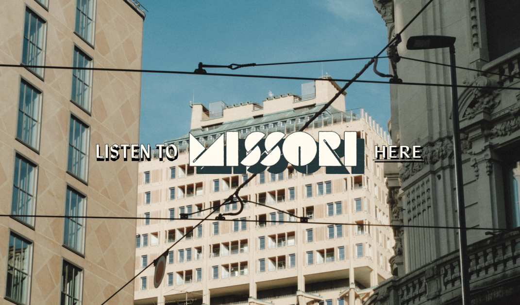 LISTEN TO MISSORI HERE - Text LISTEN TO MISSORI HERE overlaid on an urban scene with modern and historic buildings, visible windows, balconies, and overhead tram wires under a blue sky.