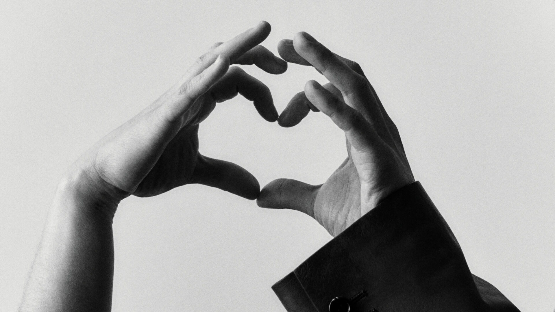 Two Hands Forming Heart - Two hands are positioned to form the shape of a heart against a plain, light background. The image is in black and white, and one hand is partially covered by a sleeve.