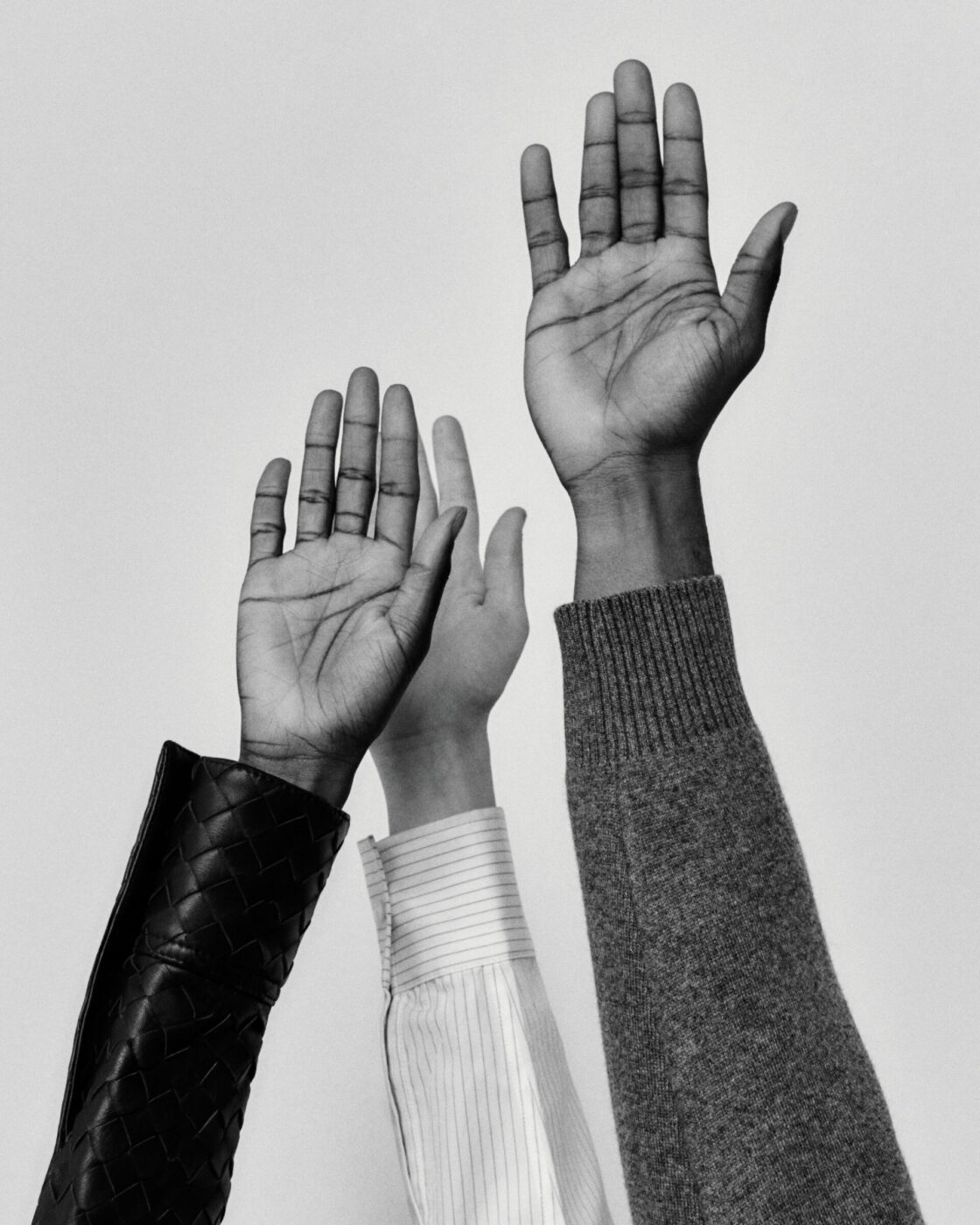 Three people raise their right hands against a plain background. Their arms show different skin tones and are dressed in different sleeves: a woven black jacket, a white striped shirt, and a gray sweater. (Note: No image title is present, so the alt text begins directly with the description.).