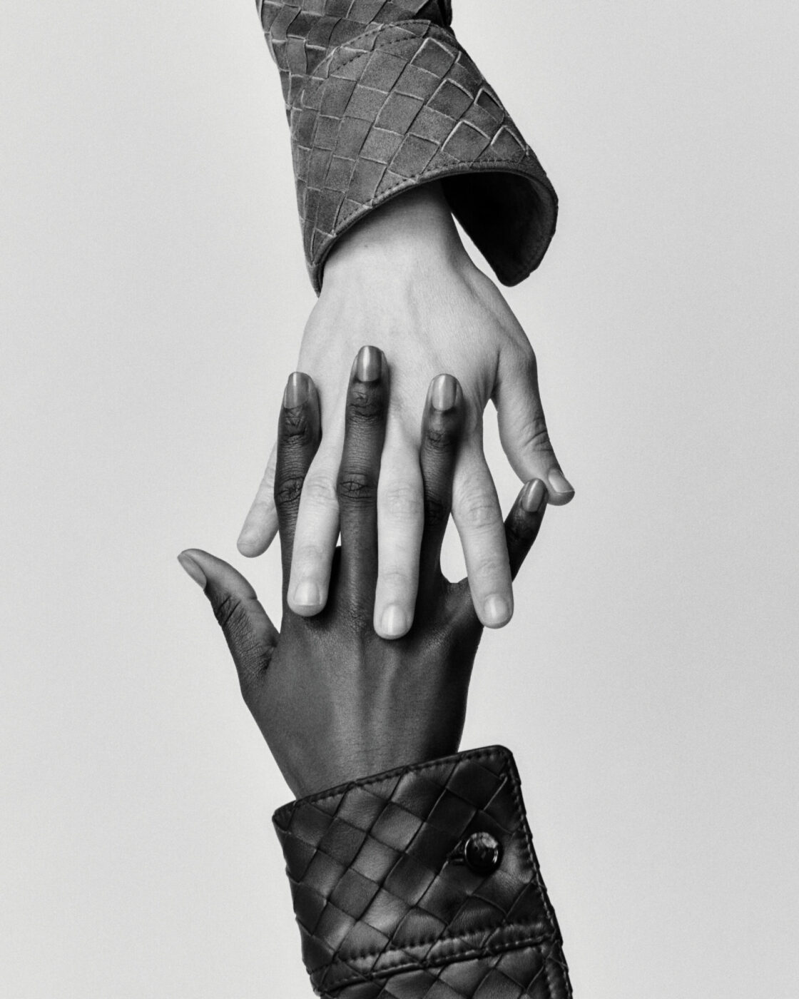 [No image title provided] - Two hands, one light-skinned and one dark-skinned, reach towards each other and touch fingers. Both wear quilted, textured jackets. The image is black and white, highlighting contrast and connection.