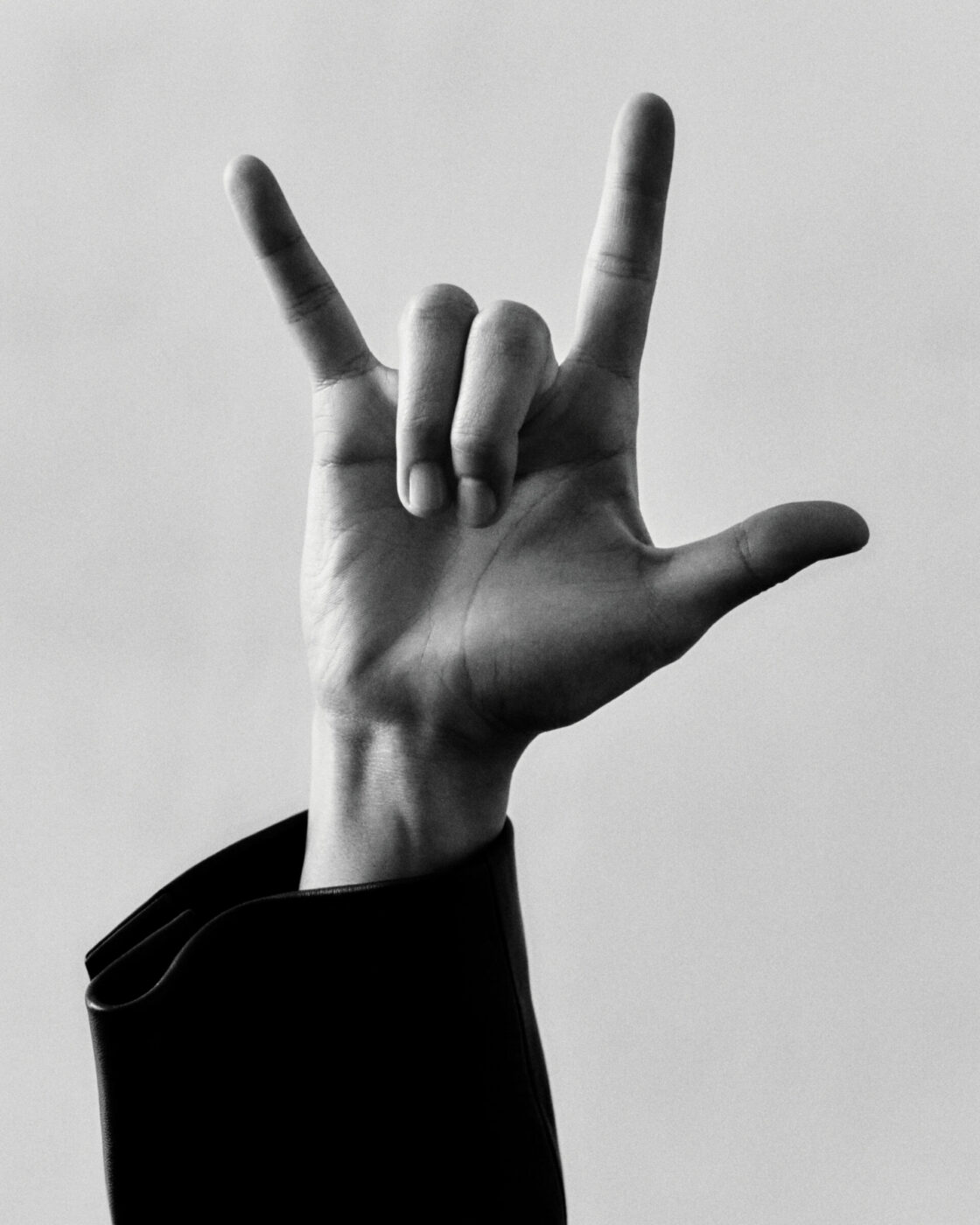 A hand in a black sleeve making the I love you gesture in American Sign Language against a plain, light background. Is this what youd like to confirm? If not, please provide more details on your request.