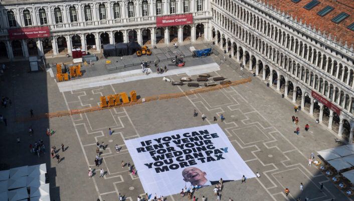 IF YOU CAN RENT VENICE FOR YOUR WEDDING, YOU CAN PAY MORE TAX - A large banner on a city square reads IF YOU CAN RENT VENICE FOR YOUR WEDDING, YOU CAN PAY MORE TAX. People walk nearby, and historic buildings surround the plaza.