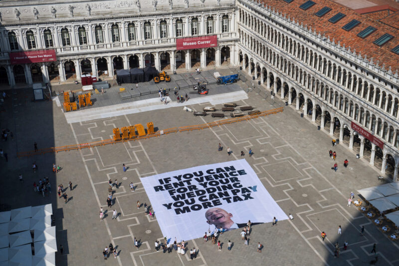 IF YOU CAN RENT VENICE FOR YOUR WEDDING, YOU CAN PAY MORE TAX - A large banner on a city square reads IF YOU CAN RENT VENICE FOR YOUR WEDDING, YOU CAN PAY MORE TAX. People walk nearby, and historic buildings surround the plaza.