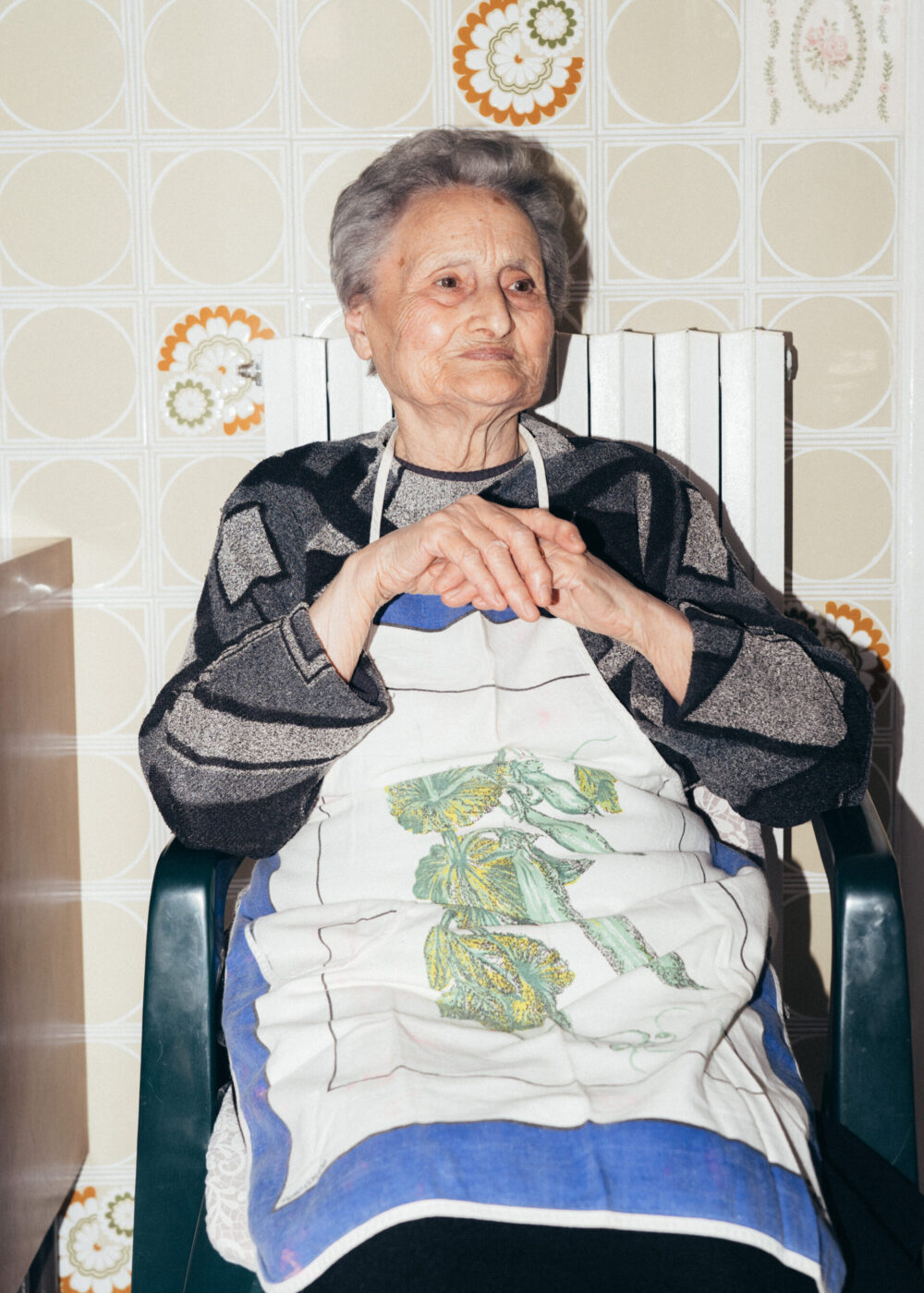 An elderly woman with short gray hair sits in a chair, wearing a patterned sweater and a white apron with a green design. She clasps her hands and looks to the side, with retro floral wallpaper in the background.