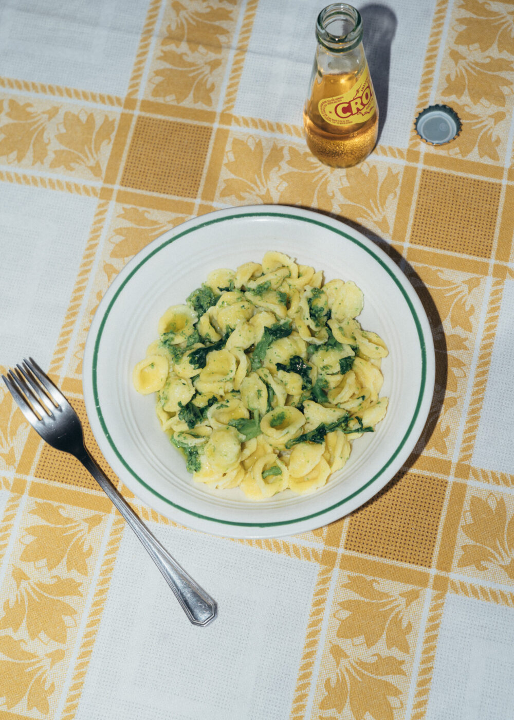 A plate of orecchiette pasta with greens sits on a yellow and white checkered tablecloth next to a fork and an open glass bottle of Crodino.