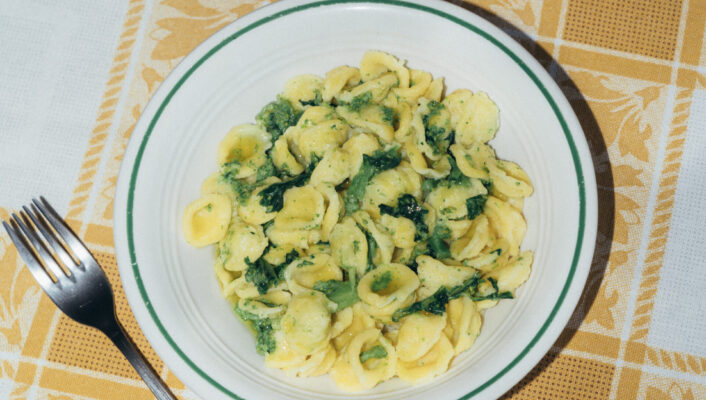 A plate of orecchiette pasta with greens sits on a yellow and white checkered tablecloth next to a fork and an open glass bottle of Crodino.