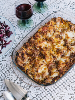 A baked lasagna in a glass dish sits on a lace tablecloth, alongside two glasses of red wine, a plate of salad, and a set of utensils.