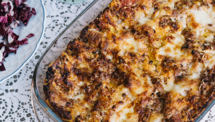 Non è stato fornito alcun titolo per l'immagine. Ecco il testo alt come da istruzioni: Una lasagna al forno in un piatto di vetro si trova su una tovaglia di pizzo accanto a due bicchieri di vino rosso, un piatto di insalata di radicchio e un set di posate.