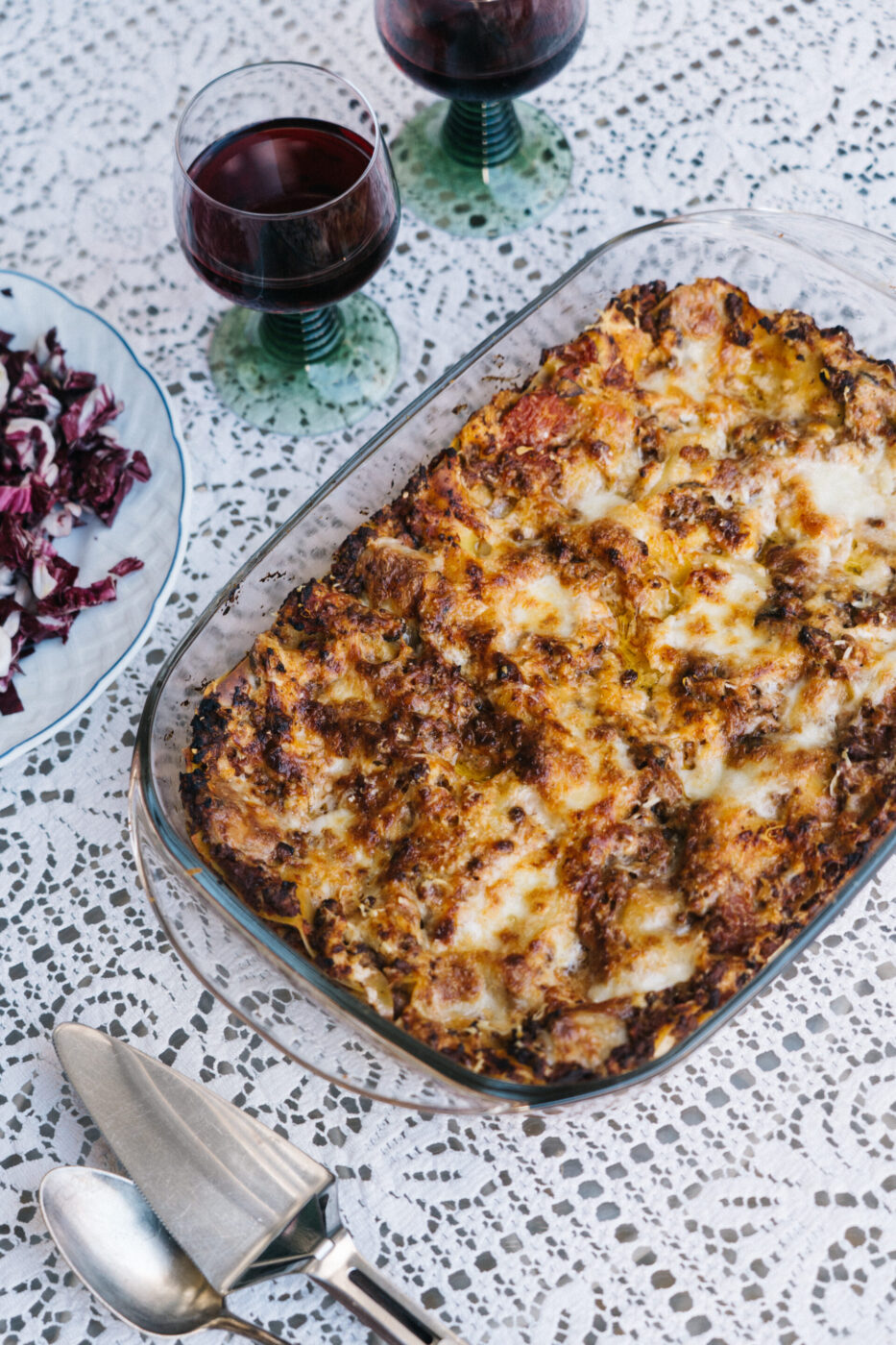 Non è stato fornito alcun titolo per l'immagine. Ecco il testo alt come da istruzioni: Una lasagna al forno in un piatto di vetro si trova su una tovaglia di pizzo accanto a due bicchieri di vino rosso, un piatto di insalata di radicchio e un set di posate.