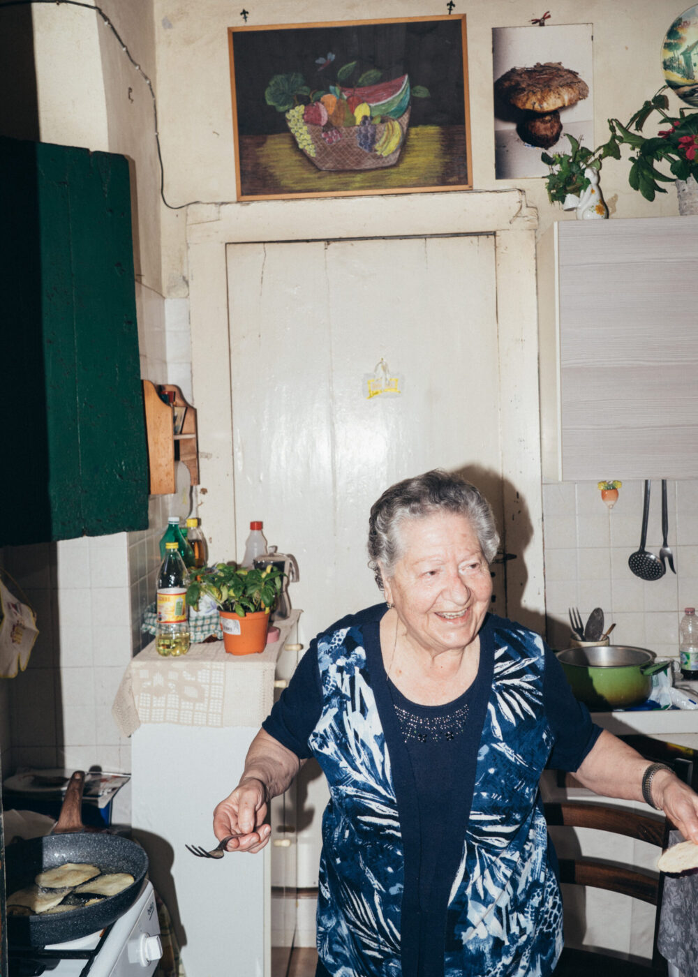 An elderly woman smiles while cooking in a small, cozy kitchen decorated with plants, artwork, and hanging utensils. She holds a fork and stands next to a stove with a frying pan. If there is no image title given, simply use: An elderly woman smiles while cooking in a small, cozy kitchen decorated with plants, artwork, and hanging utensils. She holds a fork and stands next to a stove with a frying pan. If you provide an image title, I can format it for you!.