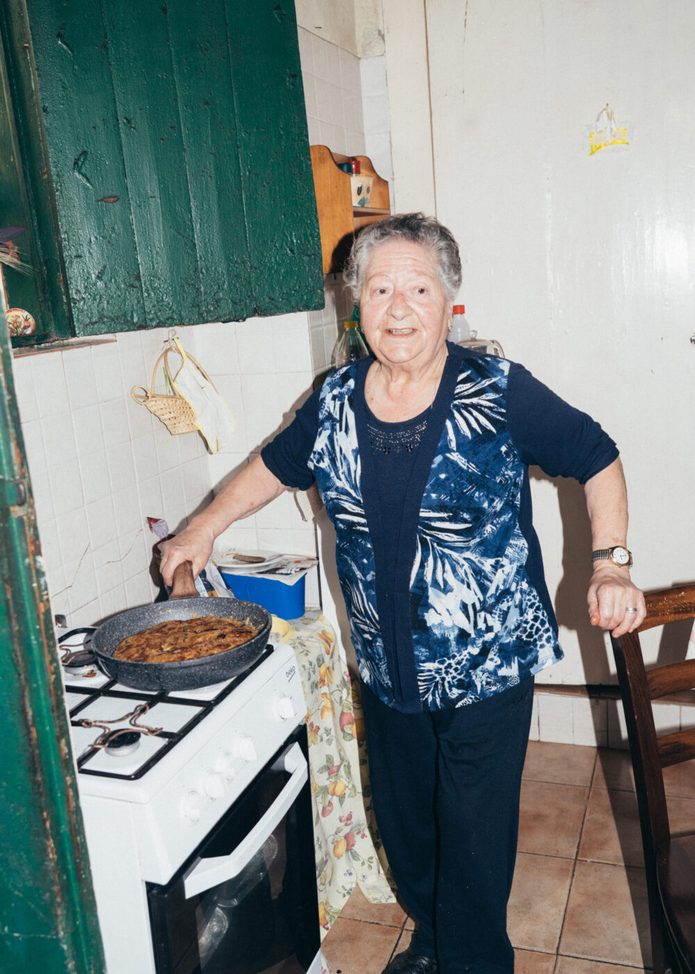Una donna anziana con i capelli corti e grigi sorride mentre cucina un grande piatto su un fornello in una cucina accogliente. Indossa un top blu a fantasia e si trova accanto a un forno bianco, con pavimenti e pareti piastrellate intorno a lei.