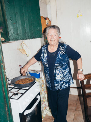 An elderly woman cooking - An elderly woman smiles while cooking a dish in a frying pan on a stove in a cozy, tiled kitchen. She wears a blue patterned vest and stands next to a green cabinet and a small table covered with a floral cloth.