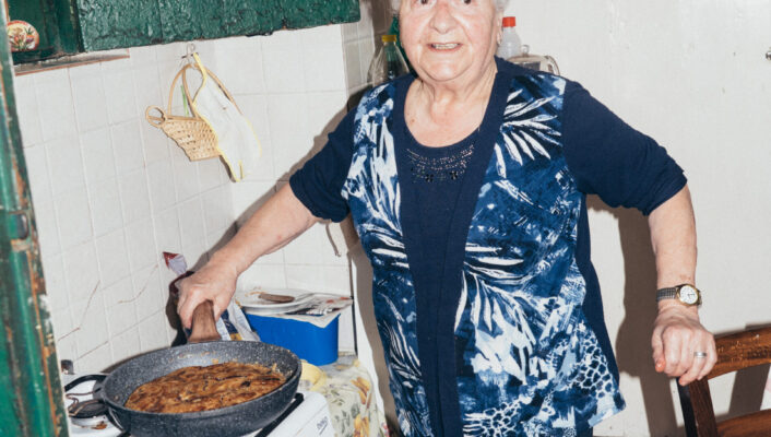 An elderly woman cooking - An elderly woman smiles while cooking a dish in a frying pan on a stove in a cozy, tiled kitchen. She wears a blue patterned vest and stands next to a green cabinet and a small table covered with a floral cloth.