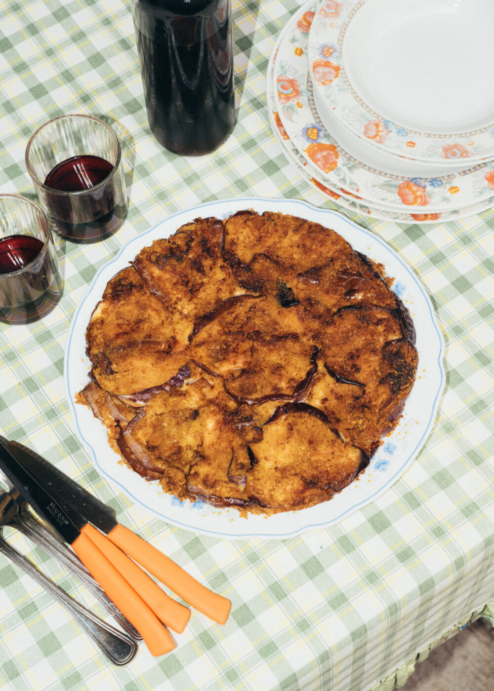 A round, golden-brown baked dish topped with slices of eggplant sits on a plate on a green plaid tablecloth, next to cutlery, orange-handled serving utensils, red wine glasses, and stacked plates.