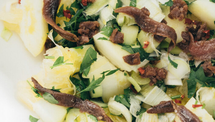 Insalata di ananas e acciughe - Primo piano di un'insalata con pezzi di ananas, sedano tritato, prezzemolo fresco, fiocchi di pepe rosso e filetti di acciuga su un piatto bianco.