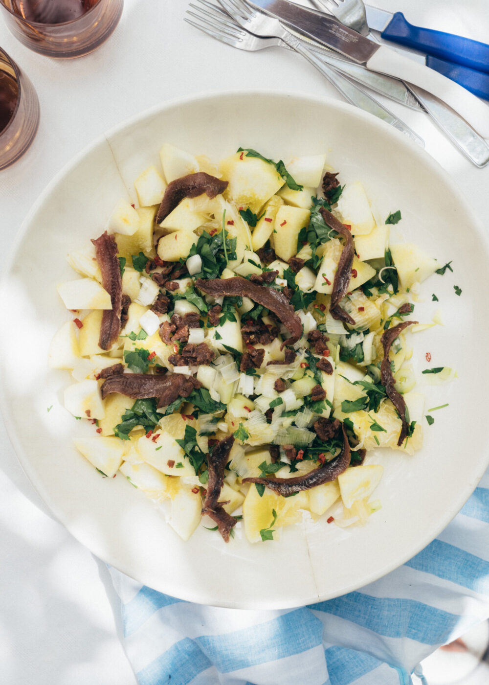 Nel vostro esempio non c'è un titolo dell'immagine, quindi il testo alt formattato sarà: Un'insalata su un piatto bianco con uova sode a pezzetti, filetti di acciuga, erbe tritate e olio d'oliva, disposta su un tavolo bianco accanto a posate e un panno a righe blu.
