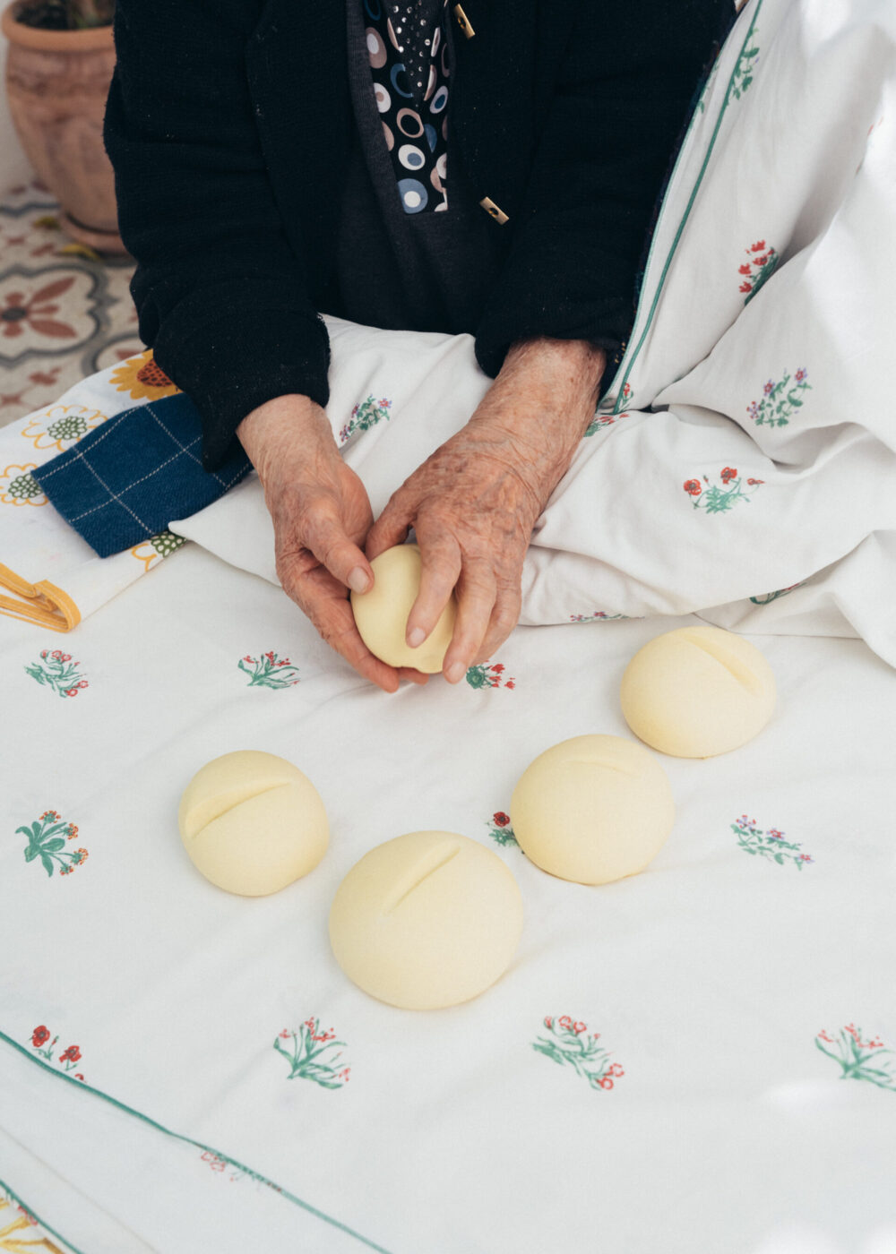 Le mani di una persona anziana modellano un pezzo di pasta rotonda su un panno bianco ornato da piccole stampe floreali. Altri cinque pezzi di pasta rotondi con fessure in cima sono appoggiati sul tessuto vicino.