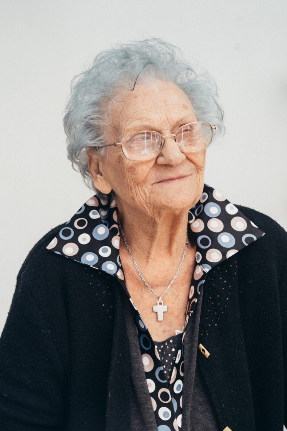 Non è stato fornito un titolo all'immagine, quindi il testo alt appropriato è: Una donna anziana con capelli grigi corti e ricci e occhiali sorride dolcemente. Indossa una giacca nera su una camicetta a pois e una collana con un ciondolo a forma di croce, su uno sfondo bianco.