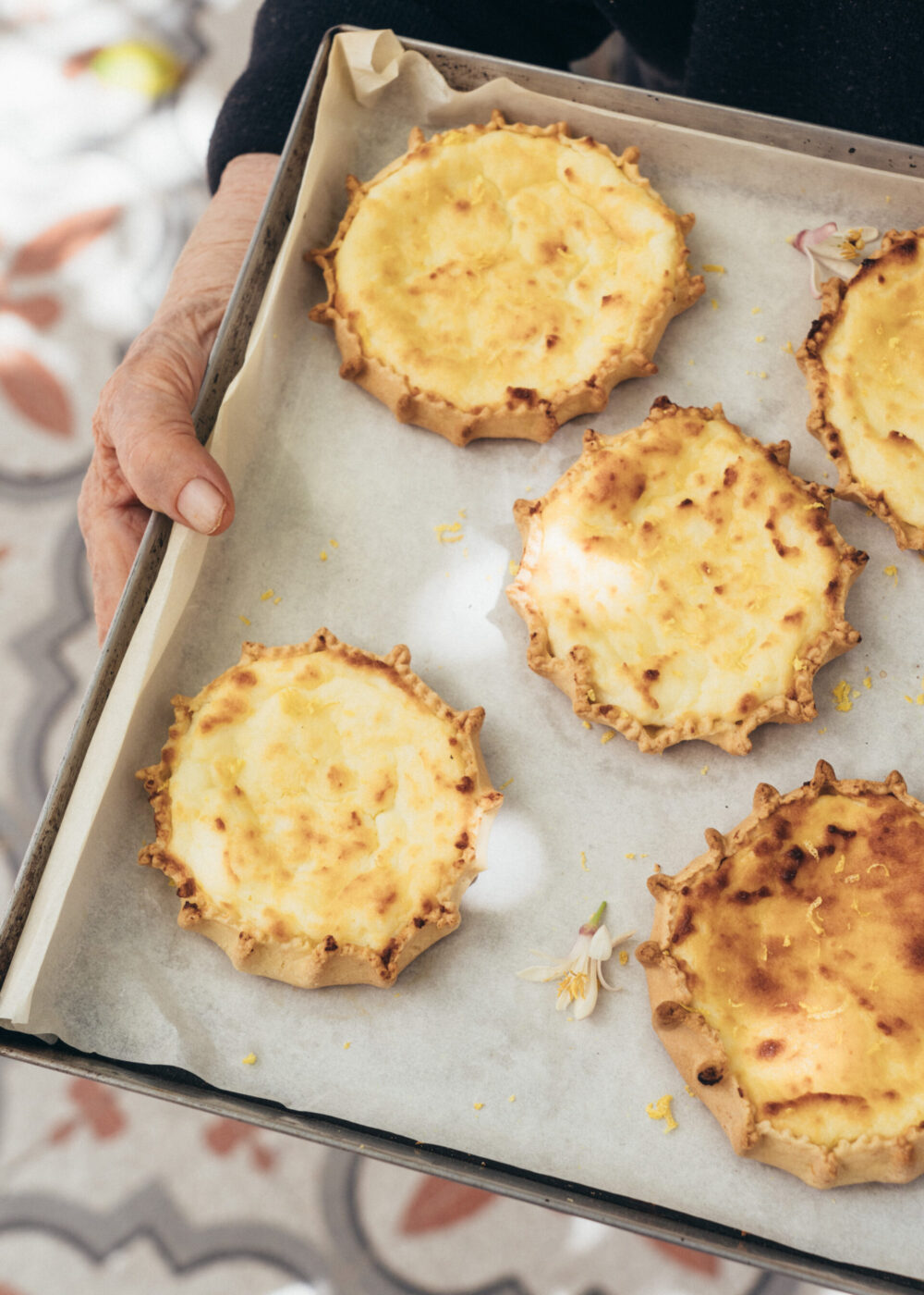 Cheese Tarts - A person holds a baking tray lined with parchment paper, displaying five golden-brown, rustic cheese tarts with crimped edges. The background shows a patterned floor.