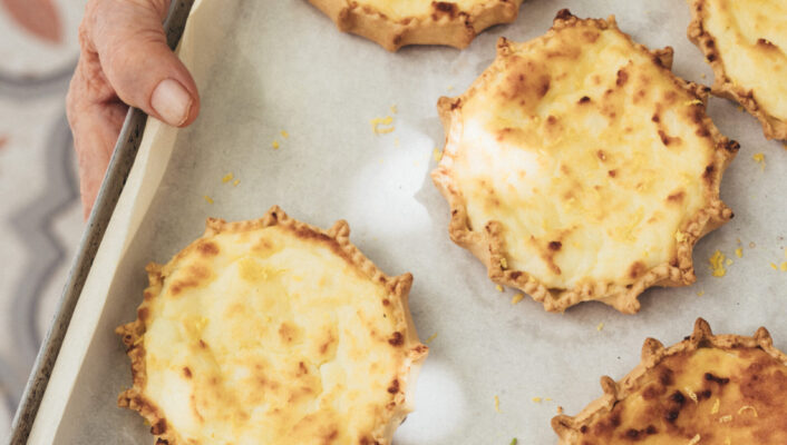 Cheese Tarts - A person holds a baking tray lined with parchment paper, displaying five golden-brown, rustic cheese tarts with crimped edges. The background shows a patterned floor.