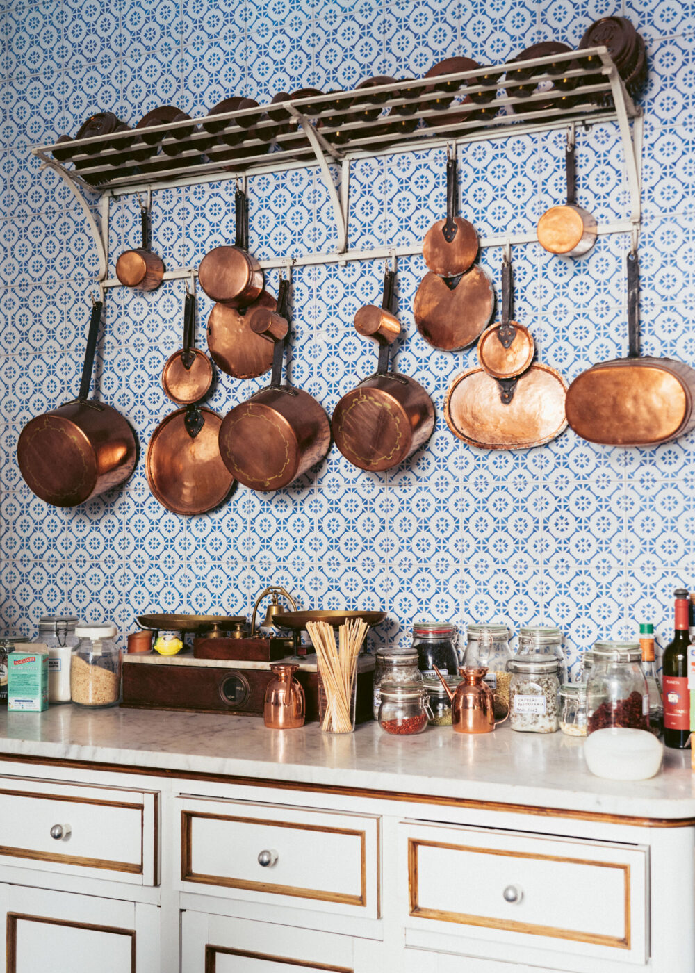 Non è stato fornito un titolo all'immagine, quindi il testo alt è: Pentole e padelle in rame sono appese a una rastrelliera sopra un bancone bianco con barattoli, spezie e utensili da cucina. Il backsplash è caratterizzato da piastrelle con motivi bianchi e blu.
